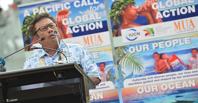 一位男士站在講臺前，其身後張貼著關於太平洋島民的海報。