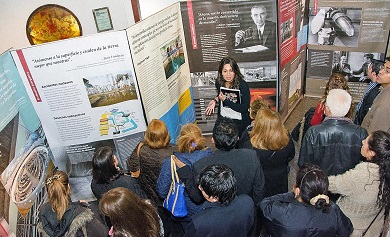 Aerial view of several people listening to a woman speaking in front of exhibition panels. 