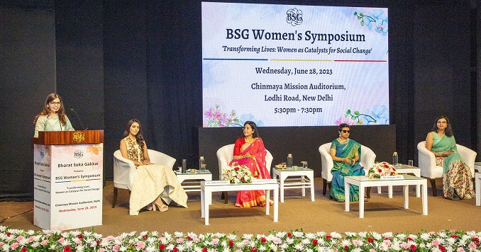 Five women on a stage, one at a podium and four seated.