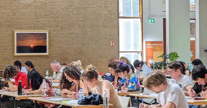 Gente en una sala sentada en pupitres realizando exámenes.