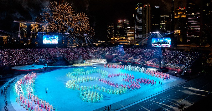 A choreographed performance at a recreation field surrounded by an audience with fireworks in the background. 