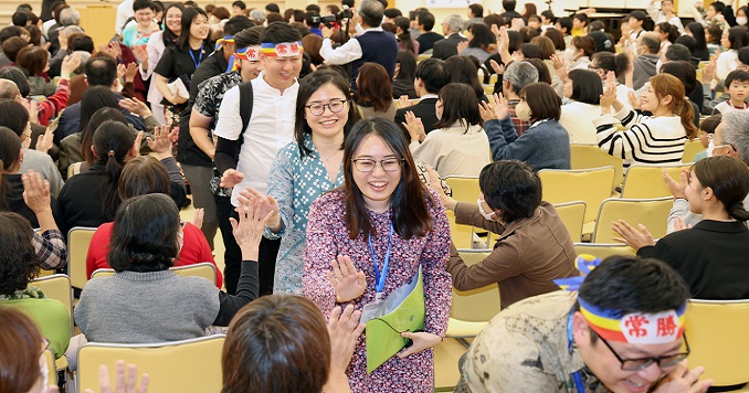 A line of people walking down an aisle exchanging greetings and farewells with those sitting in the seats lining the aisle.