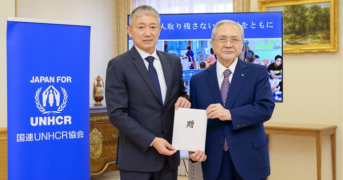 Two men holding an envelope, standing next to a Japan for UNHCR banner.