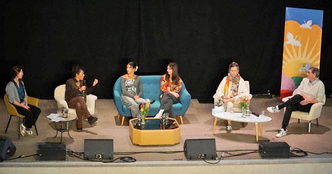 Six people seated on a stage with one person holding and speaking into a microphone.