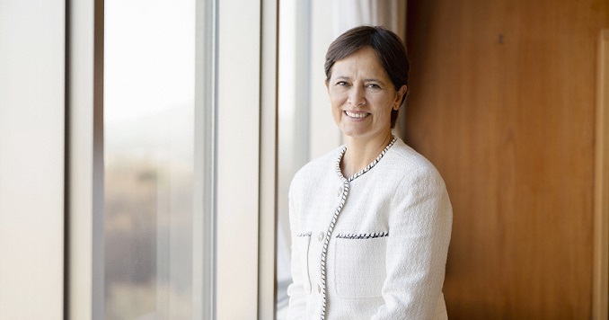 A woman smiling in a white jacket by a window.