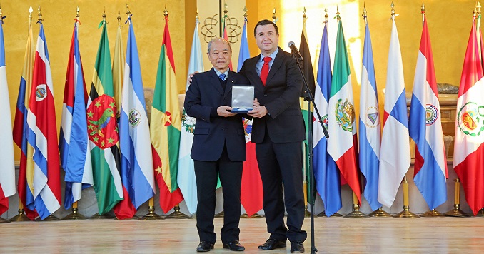 Dos hombres sosteniendo una medalla frente a unas banderas.