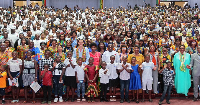 People of all ages standing in a large auditorium smiling.