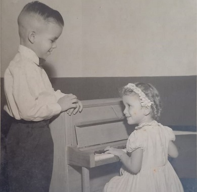 A black and white picture of a young boy looking at his sister who is playing a small piano.
