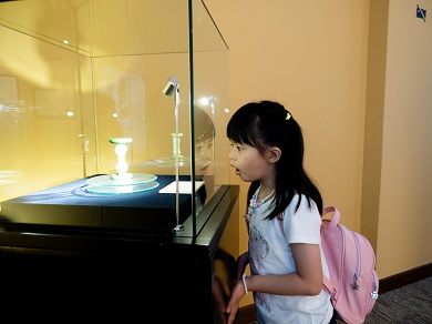 A young child wearing a backpack admires an hourglass filled with sand displayed in a glass case.