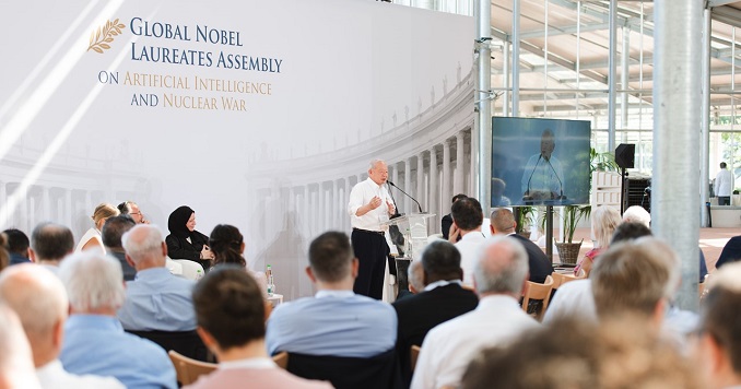 A man giving a speech, with a large conference banner behind him in a bright, glass-walled hall.