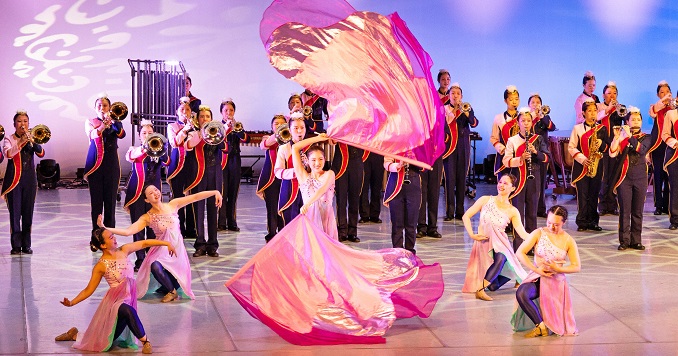 Dancers wearing long, flowing costumes perform as a marching band plays behind them.