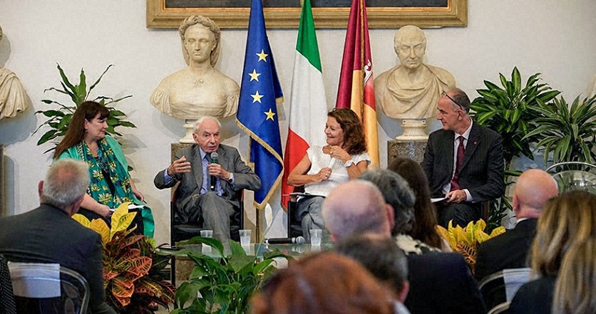Four panelists seated on stage, positioned in front of a display of three flags and two portrait busts.