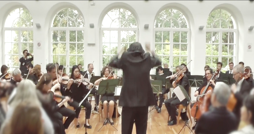 An orchestra performing in a hall with several French windows looking out onto trees
