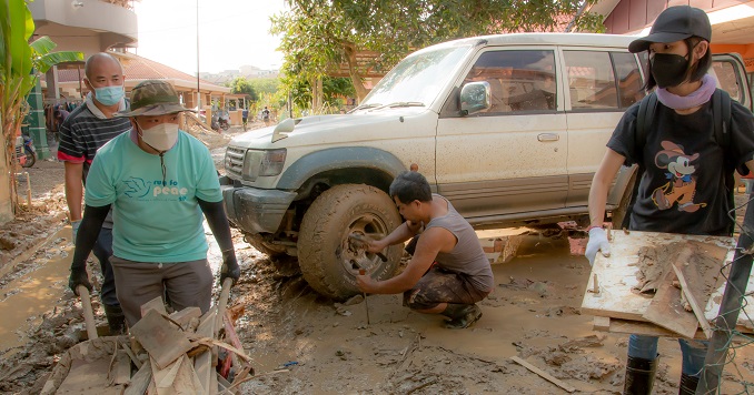 On a muddy road outside a house, a man changes a wheel on a pickup truck whilst another wheels a barrow full of broken boards.