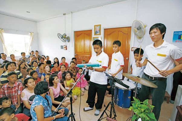 Four young people perform on musical instruments in a small room crowded with attentive listeners seated on the floor